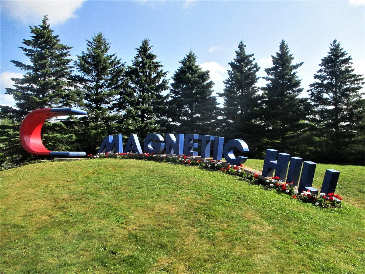 Photographie du panneau du site de la Côte magnétique à Moncton, au Nouveau-Brunswick, des lettres bleues géantes et une sculpture d'aimant en fer à cheval rouge sur une colline herbeuse